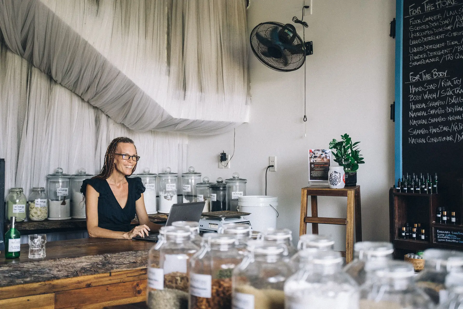 A small shop owner working on a laptop at her wooden counter, with rows of glass storage jars in the foreground